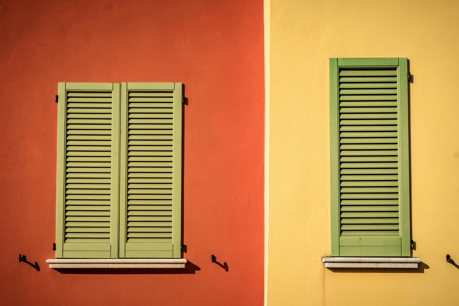 Two closed green shuttered windows on contrasting red and yellow exterior walls, showcasing different window sizes and symmetry in colorful architectural design